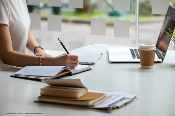 Eine Person schreibt Notizen neben einem Laptop und Arbeitsunterlagen und beschäftigt sich mit Design planen und Weiterbildung.